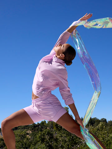 Woman in pink Tavi shorts and long-sleeve top stretching outdoors with an iridescent scarf